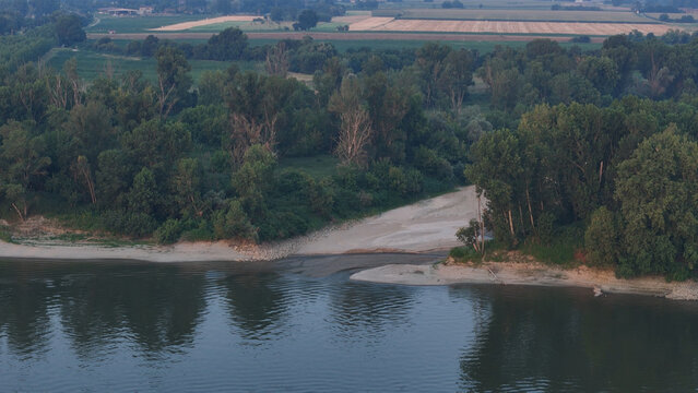 Aerial view of the Po River showing exposed sandy riverbanks due to low water levels during a heatwave, surrounded by lush green trees and agricultural fields in Cremona, Lombardy, Italy.