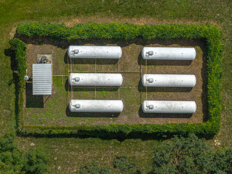 Aerial view of an LNG storage system with six white cylindrical tanks arranged in two rows within a fenced area surrounded by green hedges in Poland.