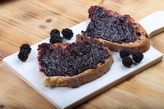 Two slices of rustic bread with thick blackberry jam and fresh berries on a white wooden board