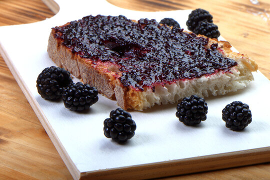 Close-up of a single slice of rustic bread with thick blackberry jam and fresh blackberries on a white wooden board