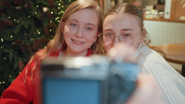 White friends review photos in cafe leaning over camera screen, soft Christmas lights and warm interior, cozy sweaters and subtle smiles, comparing shots like photography students and content