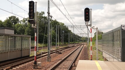 Tory kolejowe biegnące wzdłuż peronów stacji pasażerskiej pod siecią trakcyjną. Bezpieczeństwo ruchu zapewniają dwa wysokie semafory, a w tle widoczne są ekrany akustyczne oraz drzewa. © tomo81