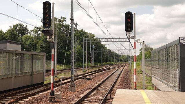 Tory kolejowe biegnące wzdłuż peron&oacute;w stacji pasażerskiej pod siecią trakcyjną. Bezpieczeństwo ruchu zapewniają dwa wysokie semafory, a w tle widoczne są ekrany akustyczne oraz drzewa.