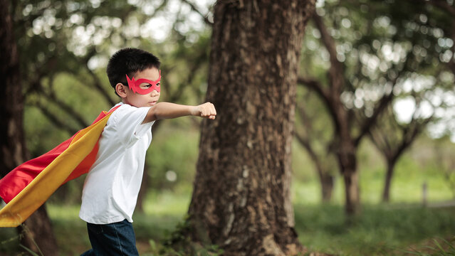 Cute Asian Boy in Superhero Costume Punching Fist Forward in Flying Pose for Child Power Concept in Green Park