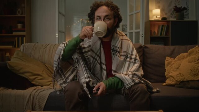 POV of middle aged Caucasian man sitting on sofa wrapped in blanket, switching channels with remote control and drinking coffee. Footage suitable for streaming service or heating ads
