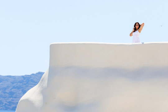 A woman in a white dress adjusts her hair on a bright white terrace overlooking the Aegean Sea in Santorini, Greece, capturing the essence of Mediterranean elegance.