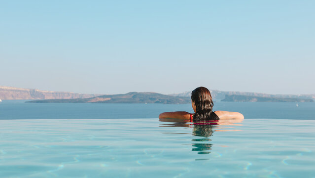 A woman floats in an infinity pool gazing toward the horizon of the Aegean Sea in Santorini, Greece, capturing the peaceful essence of Mediterranean relaxation.