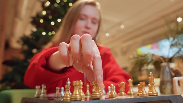 Young white woman playing chess in cafe, red sweater, focused expression and careful hand moving gold piece against blurred holiday lights and Christmas tree cozy indoor atmosphere, cinematic closeup