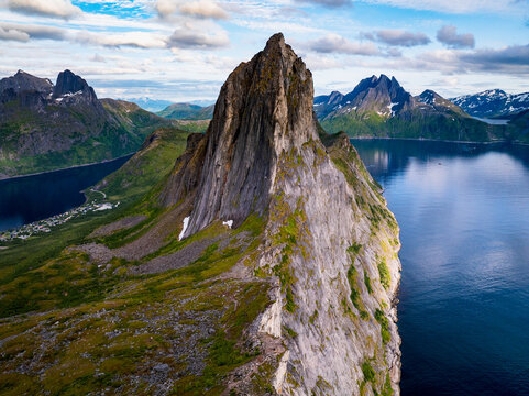 Aerial view of Segla mountain peak rising above the deep blue waters of the fjord with rugged cliffs, green slopes, and distant snow-capped mountains in Senja, Troms, Norway.