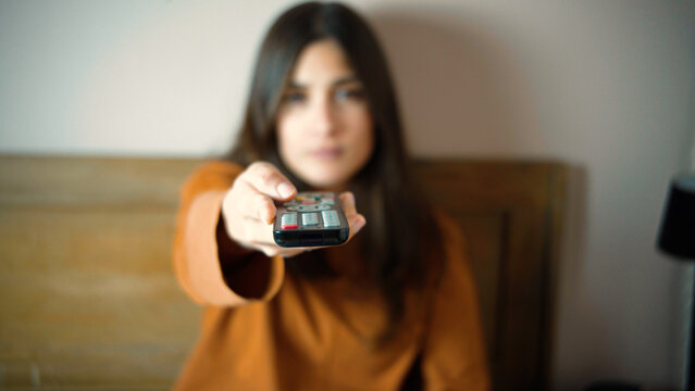 Close-up of a young Hispanic woman's hand while she sits in bed, changing channels with her TV remote, representing entertainment, relaxation, leisure, and everyday home life.