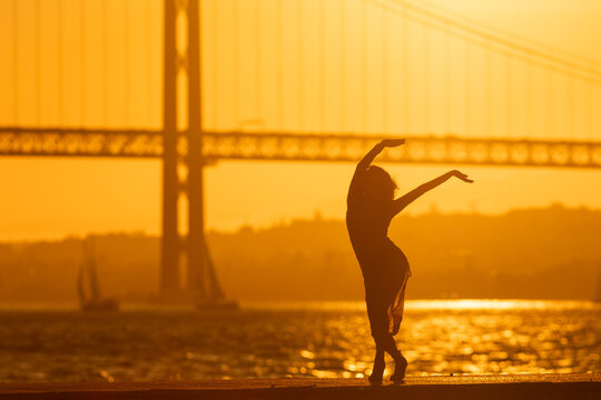 Dancer in silhouette poses by the water during sunset near the bridge