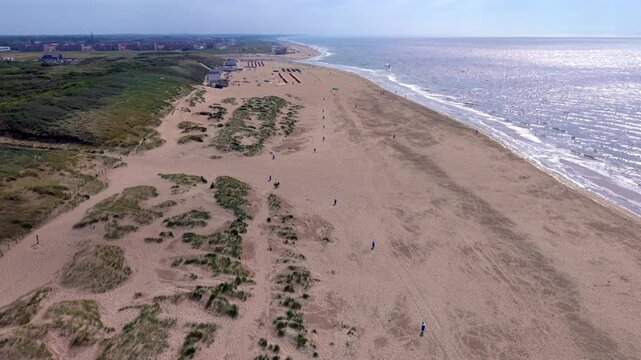 Aerial view video clip of Katwijk aan Zee wide sandy beach, coastal sand dunes, beach cabins, and sunlit North Sea waves in South Holland, Netherlands on a bright summer day