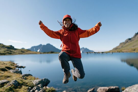 Joyful hiker jumping by mountain lake in red jacket with backpack for outdoor adventure and travel background