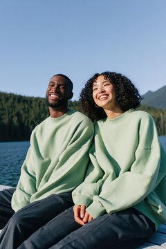 Smiling multicultural couple in oversized sage sweatshirts on a lakeside pier for relaxed lifestyle background and outdoor banner design