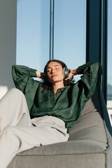 Fototapeta premium Person relaxing with headphones on a gray lounge chair by a large window in sunlit modern interior for mindfulness background and lifestyle banner