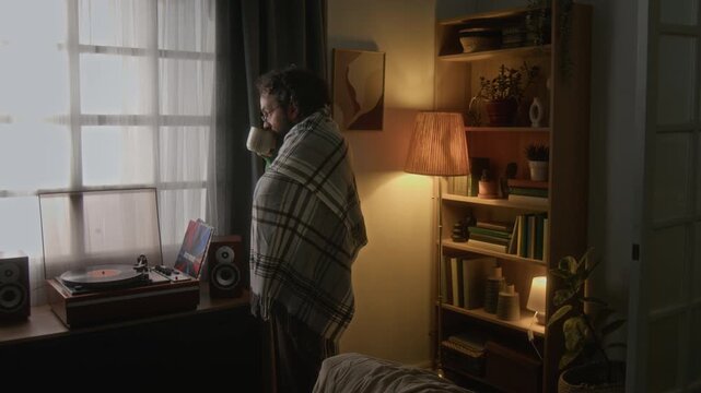 Middle-aged Caucasian man wrapped in blanket standing by window, drinking hot beverage and looking outside. Video is perfect for mental health, wellness, and self-care marketing campaigns