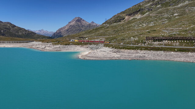 Aerial view of the red Bernina Express train traveling along the turquoise waters of Lago Bianco near the Bernina Pass in Poschiavo, Grisons, Switzerland.