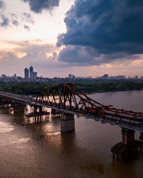 Aerial view of the historic Long Bien Bridge crossing the Red River with a train passing over, set against a dramatic sunset sky and the city skyline in Hong Ha, Ha Noi, Vietnam.