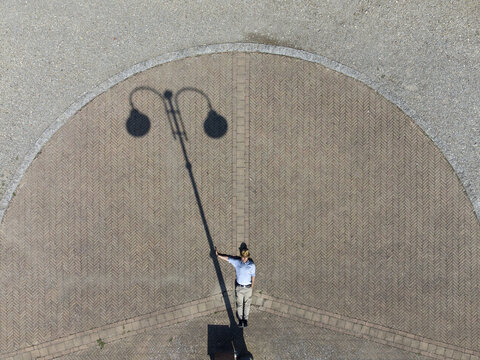 Aerial view of a person standing on a paved area with a long shadow cast by a street lamp in Alessandria, Piedmont, Italy.