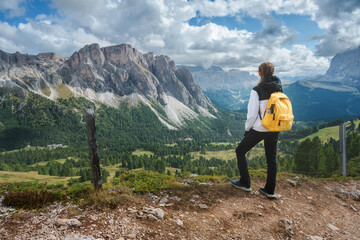 Obraz premium Woman hiker with yellow backpack standing on trail looking at mountain range in Dolomites, Italy. Alpine summer landscape with female tourist enjoying valley view, rocky peaks and cloudy sky. Hiking