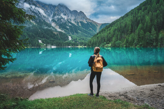 Woman hiker with yellow backpack standing near turquoise mountain lake in Dolomites, Italy. Alpine summer landscape with crystal clear water reflection, pine forest, rocky peaks, clouds in rainy day