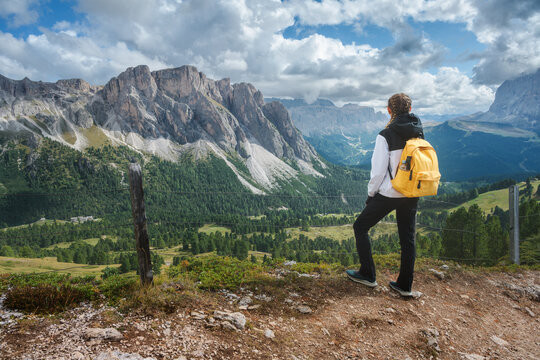 Woman hiker with yellow backpack standing on trail looking at mountain range in Dolomites, Italy. Alpine summer landscape with female tourist enjoying valley view, rocky peaks and cloudy sky. Hiking