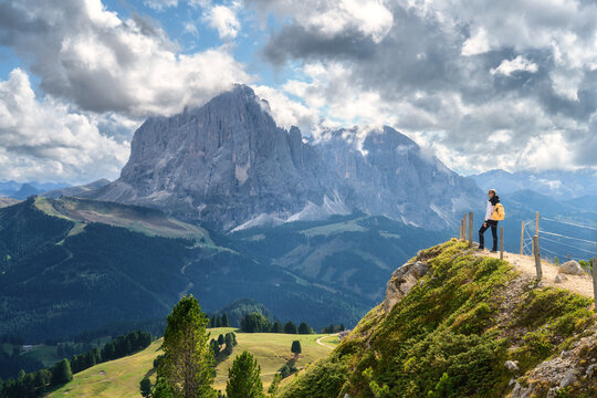 Woman hiker with backpack standing on cliff edge looking at mountain peak. Dolomites, Italy. Alpine summer landscape with female tourist on view point, green valley, rocks, cloudy sky. Hiking in Alps