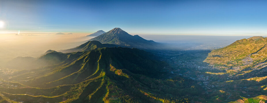 Aerial view of the volcanic landscape of Dieng Plateau with lush green mountains, terraced agricultural fields, and distant peaks under a clear morning sky in Indonesia.