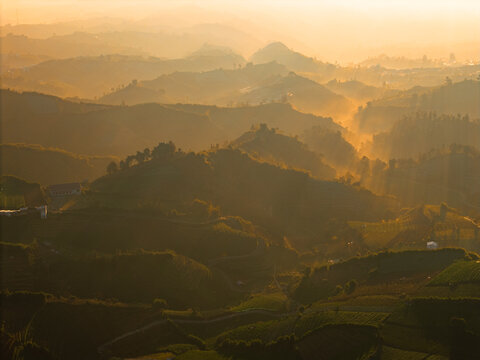 Aerial view of Dieng Plateau at sunrise, with rolling mountains, lush agricultural terraces, and golden light filtering through the morning mist in Indonesia.