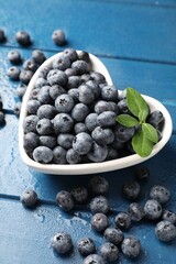 Fresh ripe blueberries with green leaves in heart shaped bowl on blue wooden table, closeup © New Africa