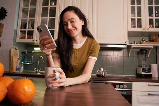 Woman smiles at phone while holding a mug in kitchen