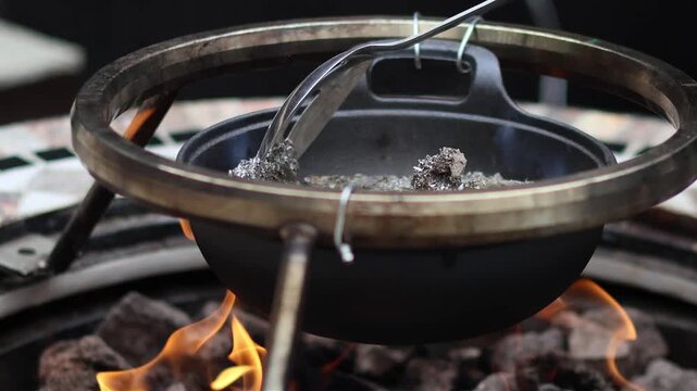 Close up of removing dross impurities from molten metal scrap in cast iron pot over open flame. Preparing clean melted lead or tin to cast handmade items, fishing weights and sinkers in home workshop.