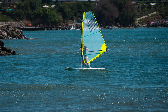 A person enjoys a sunny summer day windsurfing on bright blue water with a yellow-edged transparent sail near a rocky, tree-lined coast.