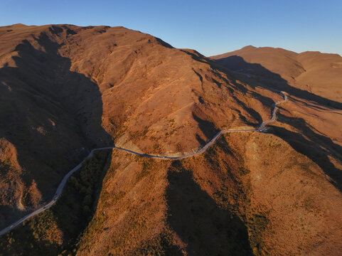 Aerial view of the winding Crown Range Road cutting through the rugged, sunlit mountains at Roaring Meg, Otago Region, New Zealand.