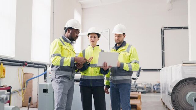 Group of three factory engineers wearing safety jackets and helmets discussing project. Female holding laptop showing data while colleague using tablet. Team discussing factory details.