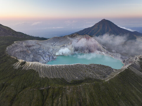 Aerial view of the Ijen volcano crater featuring its iconic turquoise acidic lake, rugged rocky rim, and sulfurous steam vents set against a mountainous landscape in East Java, Indonesia.