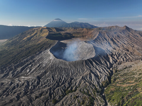 Aerial view of Mount Bromo with its volcanic crater emitting smoke, surrounded by rugged terrain and distant mountains in Tosari, East Java, Indonesia.