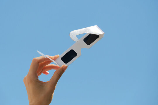 Woman hand holds solar eclipse glasses against blue sky background. Solar eclipse glasses are specially certified, ultra-dark glasses to protect eyes from damage when looking directly at sun.