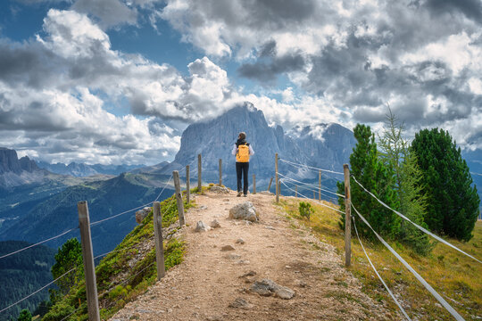 Woman hiker with yellow backpack walking along dirt trail towards rocky mountain peak in Dolomites, Italy. Alpine summer landscape with female tourist on gravel path under dramatic cloudy sky. Hiking