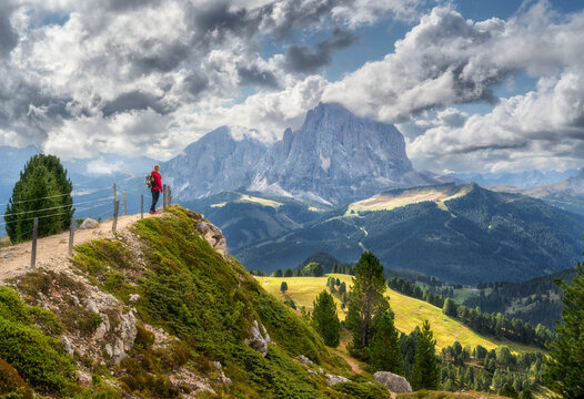 Woman hiker in red jacket with backpack standing on cliff edge viewpoint looking at mountain peak in Dolomites, Italy. Alpine summer landscape with green valley, trail, fence and dramatic cloudy sky