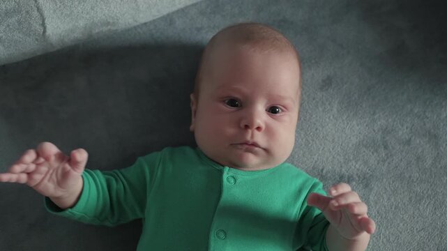Newborn rests on textured gray blanket while wearing green onesie, both arms lifted in visible reaching motion, fingers splayed, head turned slightly, eyes open, calm focused face