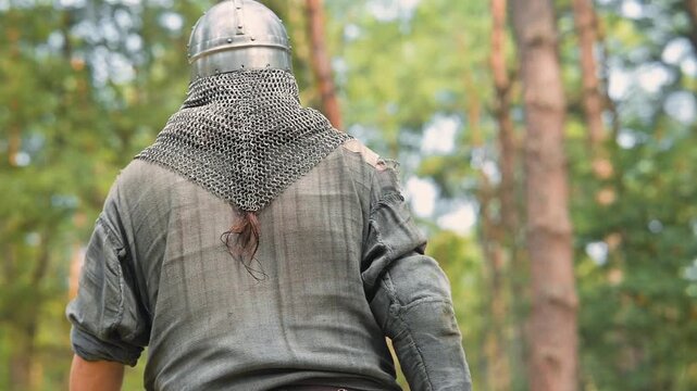 A man in a chainmail coif moves through a wooded area, wearing a simple tunic and belt. This scene captures a moment before battle in a historical reenactment of the early Middle Ages