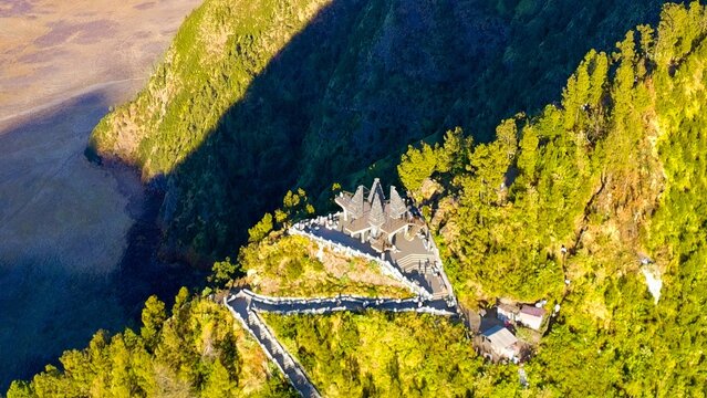 Aerial view of Seruni Point viewing platform perched on a steep cliff edge surrounded by lush green pine trees and rugged mountain terrain in Kecamatan Puspo, Jawa Timur, Indonesia.