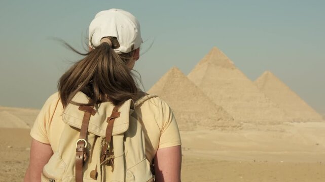 Caucasian woman tourist with backpack walking towards pyramids in Giza on sandy hill clear sunny day, visitor exploring sights of ancient Egypt in Cairo