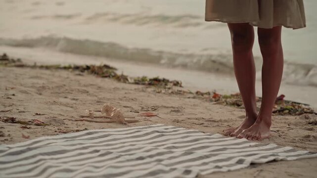 Striped towel tote on beach shore, soft dawn light, bare feet stepping on sand, woman spreading striped blanket, placing woven bag on blanket, gentle waves lapping, scattered seaweed on shore, calm