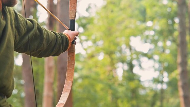 An archer prepares to shoot an arrow in a dense forest. The person is dressed in simple medieval clothing and holds a wooden bow, focusing on their target