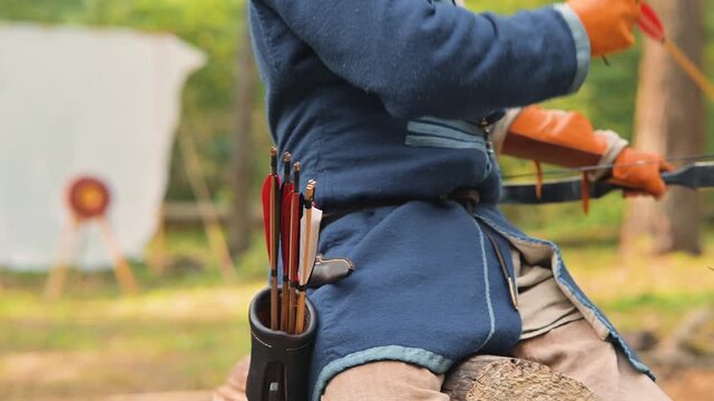 An archer prepares to shoot arrows towards targets placed in a forest clearing. The archer is dressed in a blue tunic, with a quiver of arrows at their side, set against a backdrop of trees