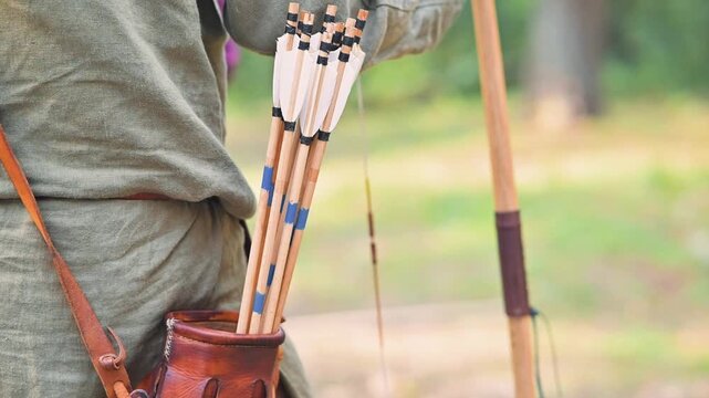 An individual stands in a forest, wearing a simple shirt and leather accessories, reaching for arrows in a quiver