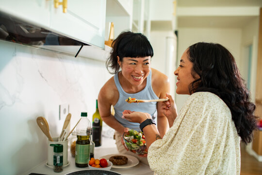 Two women cooking and tasting salad in a home kitchen