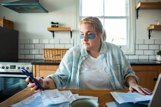 Woman with under-eye patches paying bills at kitchen table